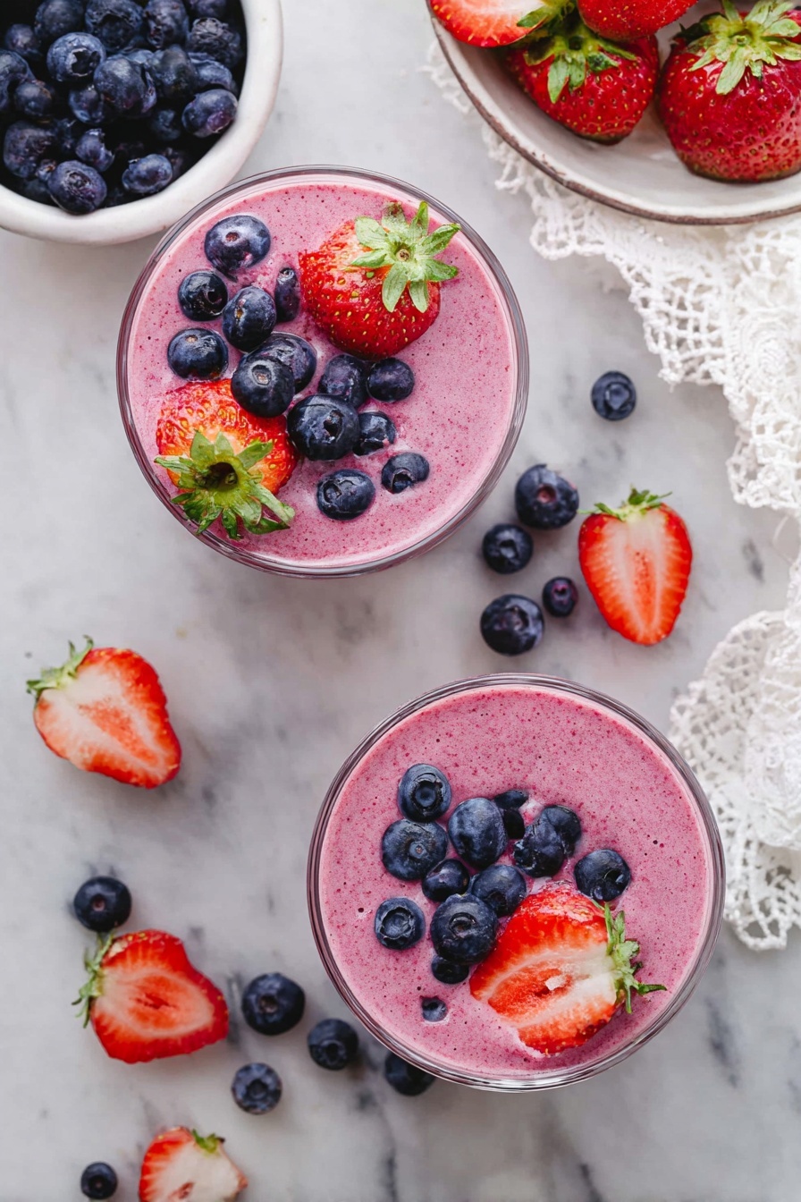 Two clear glass cups filled with a pink mixed berry smoothie, both topped with fresh blueberries and halved strawberries with green leaves. Around the cups, there are scattered blueberries and halved strawberries on a white marbled surface. Nearby, one white bowl is filled with blueberries and another white plate holds whole strawberries and blueberries. A piece of white lace cloth is placed in the upper right corner. photo taken with an iphone --ar 2:3 --v 7