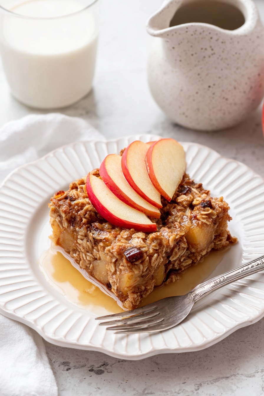 A square piece of baked oatmeal sits on a white plate with scalloped edges, showing a textured mix of oats, small apple chunks, and raisins in warm brown and beige tones. The top layer is decorated with three thin apple slices with red edges and pale yellow centers, arranged in a slight fan shape. The oatmeal piece is glossy with syrup pooled around its base on the plate. A silver fork rests on the plate beside the oatmeal. In the background, there is a white marbled surface with a white speckled ceramic pitcher and a glass of milk. photo taken with an iphone --ar 2:3 --v 7