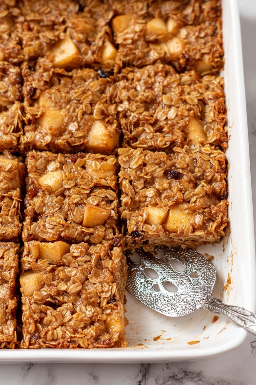 The image shows a close-up of a white baking dish filled with a baked apple and oat casserole cut into six square pieces. The top layer is golden brown and uneven, with visible chunks of cooked apples scattered throughout. The texture looks moist and crumbly with oats and raisins integrated into the mix. To the right side inside the dish, a silver serving spatula with decorative cut-outs lies flat, slightly under one piece. The dish sits on a white marbled surface. photo taken with an iphone --ar 2:3 --v 7
