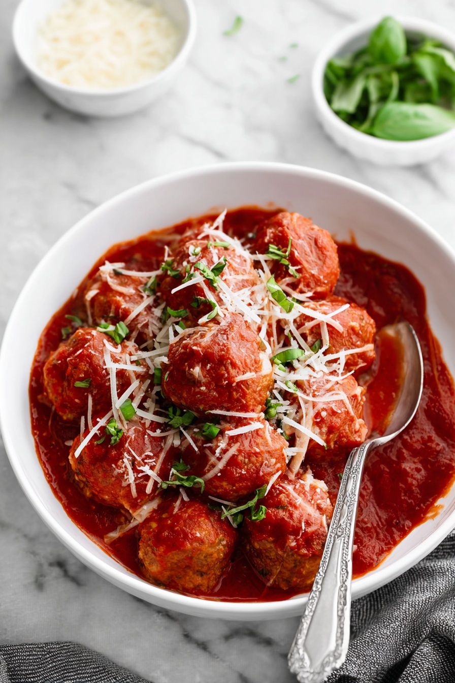 A white bowl filled with round meatballs covered in thick red tomato sauce, topped with shredded white cheese and small green basil pieces. The meatballs are arranged closely, almost filling the bowl, with sauce pooling around them. A silver spoon with a detailed handle rests inside the bowl on the right side. The bowl sits on a white marbled surface, and in the background, there is a small white bowl with fresh green basil leaves and another white bowl with grated cheese. photo taken with an iphone --ar 2:3 --v 7