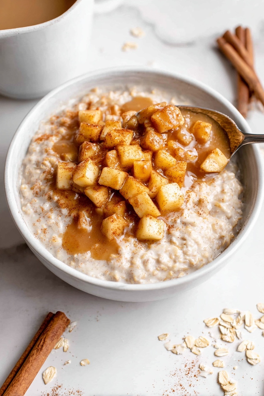 A white bowl filled with creamy oatmeal as the base layer, light beige in color and soft texture. On top, there is a thick layer of small, cubed cooked apples, golden brown with a sprinkle of cinnamon giving a warm, spicy look. The oatmeal and apple cubes are mixed with a drizzle of light brown sauce, pooling slightly around the sides. A metal spoon is scooping some of the oatmeal and apples from the bowl. The bowl is placed on a white marbled surface with a few cinnamon sticks and scattered oats nearby. Photo taken with an iphone --ar 2:3 --v 7
