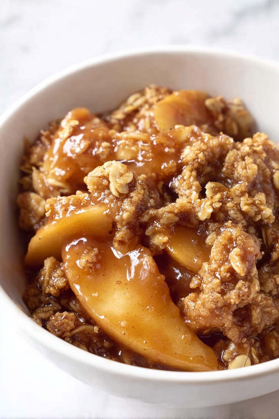 The image shows a close-up of a white bowl filled with a warm apple crumble dessert. The bottom layer consists of thick, soft apple slices coated in a shiny, sticky caramel-like sauce with a rich brown color. On top, there is one thick layer of crumbly, textured oat and brown sugar topping that looks slightly toasted and golden, adding a rough and crunchy contrast to the soft apples underneath. The dessert appears moist and inviting with the sauce glistening in the light. The bowl sits on a white marbled surface. Photo taken with an iphone --ar 2:3 --v 7