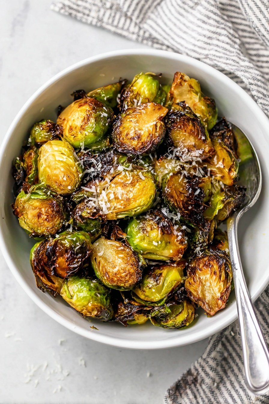 A white bowl filled with roasted Brussels sprouts that are golden brown and slightly charred on the edges, showing a mix of crispy dark green outer leaves and softer caramelized yellow-green inner layers. The Brussels sprouts are mostly halved, exposing their round, layered texture with a light grating of white cheese sprinkled on top. A silver spoon rests inside the bowl on the right side. The bowl is placed on a white marbled surface with a folded striped cloth napkin in the background. photo taken with an iphone --ar 2:3 --v 7