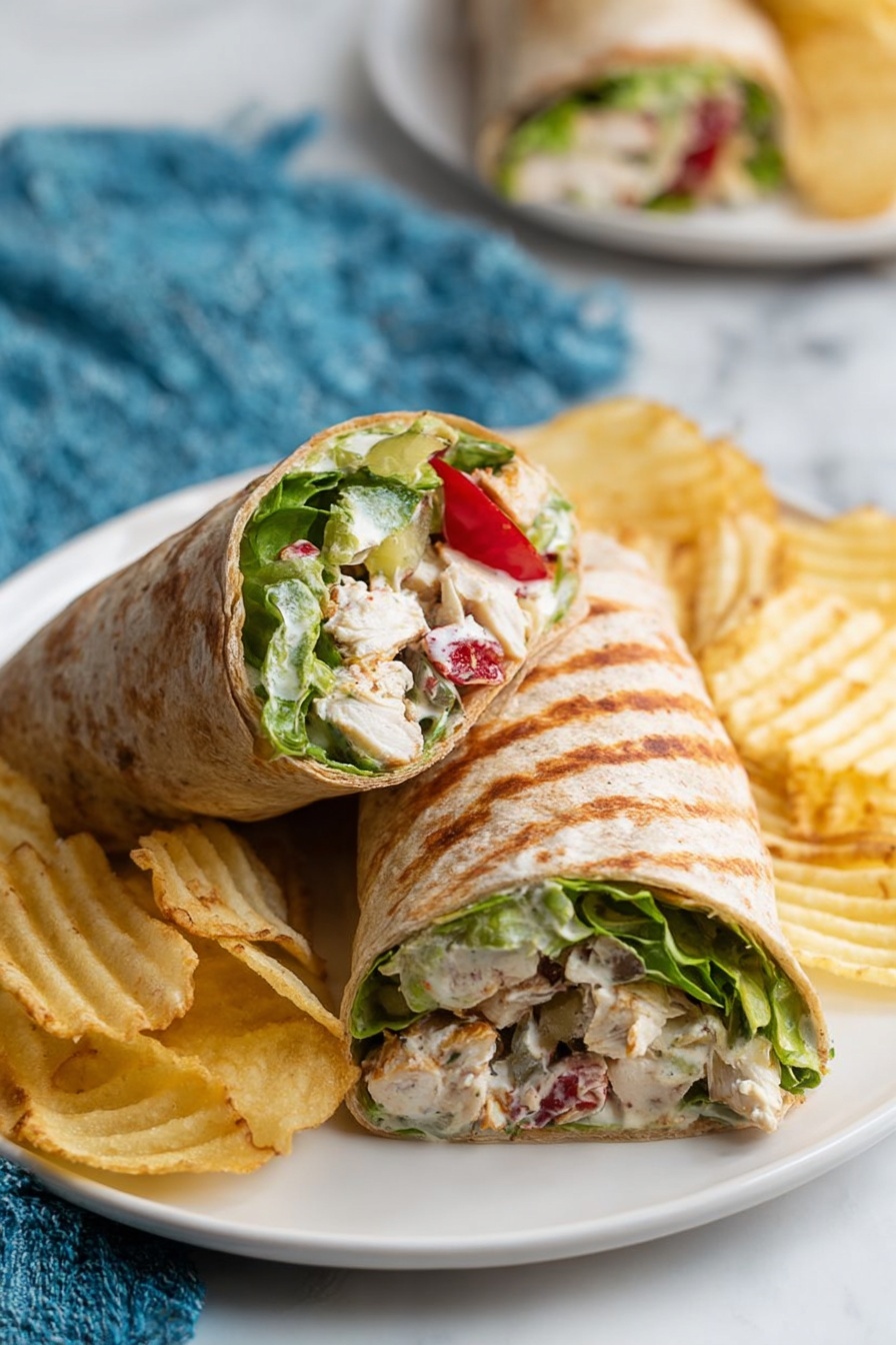 A white plate on a white marbled surface holds two halves of a wrap sandwich. The wrap is light brown with darker toasted spots and filled with layers of green lettuce, chunks of white chicken, and small red tomato pieces mixed with a creamy white sauce. Several crinkly, pale yellow ridged potato chips are scattered around the wrap. In the background, there is a blurred look of another plate with similar wraps and potato chips. A blue fabric is partially seen near the plate. photo taken with an iphone --ar 2:3 --v 7