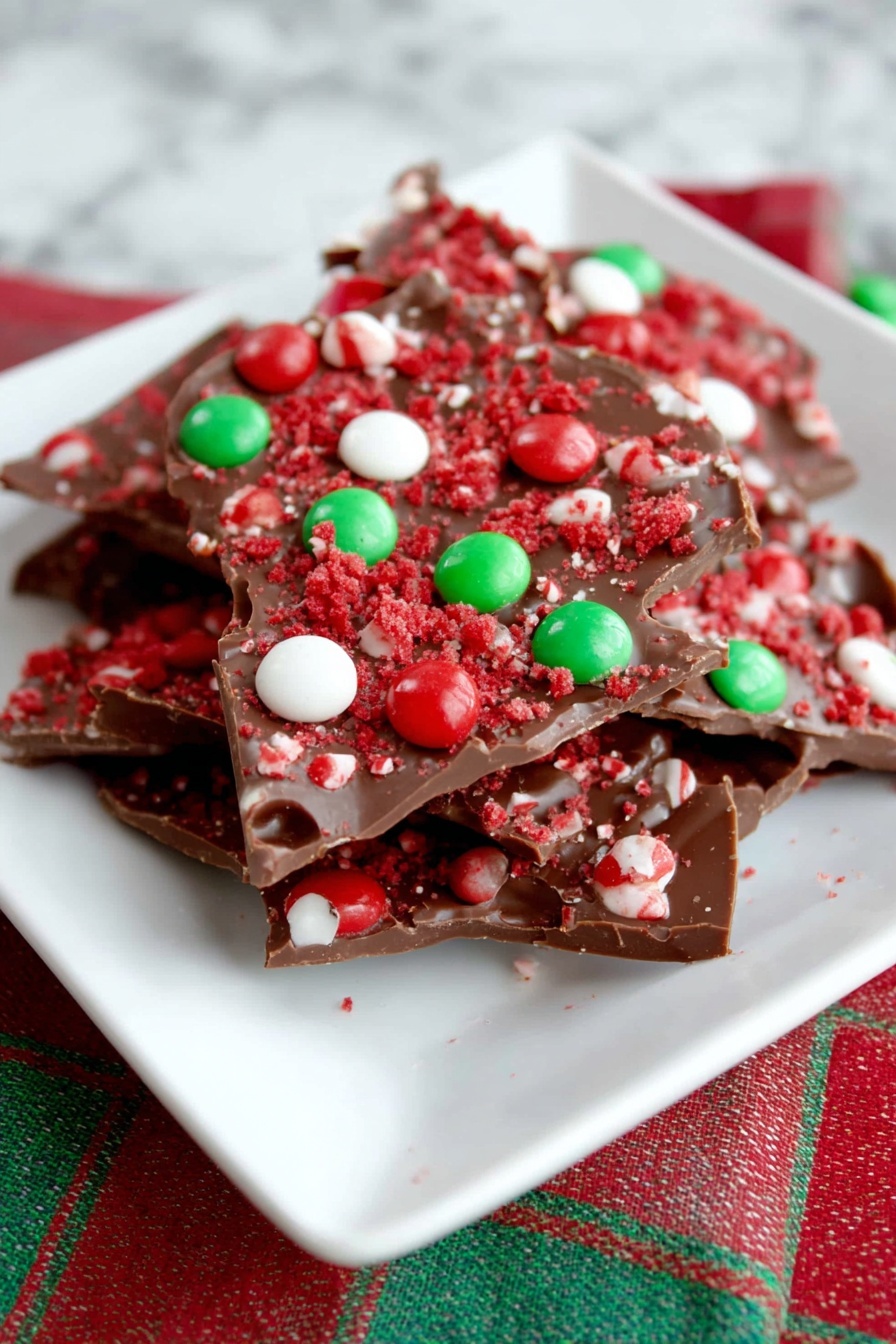 A white square plate holds several stacked pieces of chocolate bark. Each piece is dark brown and topped with bright red crumbs and scattered round candies in red, white, and green colors, creating a festive look. The bark pieces have rough edges and some holes in the chocolate. The plate sits on a white marbled surface with a red and green striped cloth partially visible beneath. photo taken with an iphone --ar 2:3 --v 7
