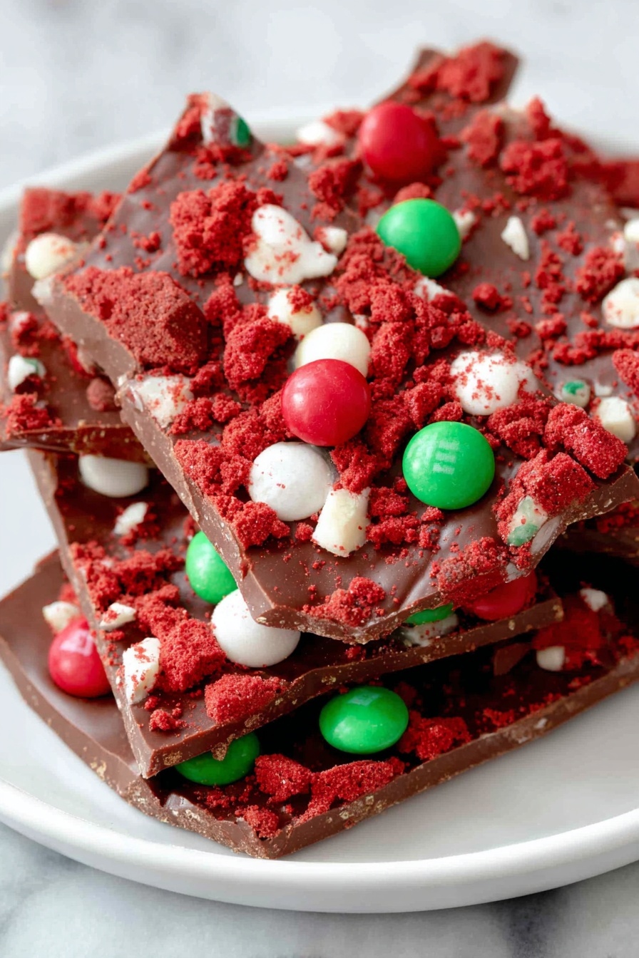 The image shows a stack of broken chocolate bark pieces on a white plate. The chocolate bark has a smooth, rich brown base layer topped with crumbled red cake pieces giving a rough, crumbly texture. Scattered on top are round candy-coated chocolates in red, white, and green colors adding bright, glossy spots. The bark pieces are unevenly layered with some pieces resting on others, showing depth and a mix of textures from smooth chocolate to crumbly cake and shiny candies. The plate rests on a white marbled textured surface. Photo taken with an iphone --ar 2:3 --v 7