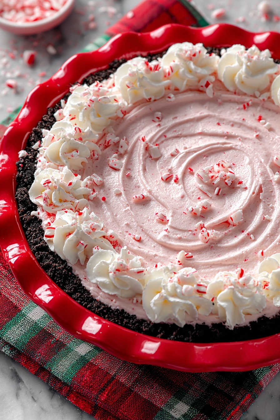 The image shows a dessert with three main layers in a white pie dish with a red rim. The bottom layer is a dark crumbly crust that looks like crushed cookies. On top of that is a thick, smooth light pink cream spread evenly in a swirl pattern. Around the edge, there are dollops of white whipped cream. The entire dessert is sprinkled with small pieces of crushed red and white candy. The background is a white marbled surface with a blurred Christmas tree and ornaments. Photo taken with an iphone --ar 2:3 --v 7
