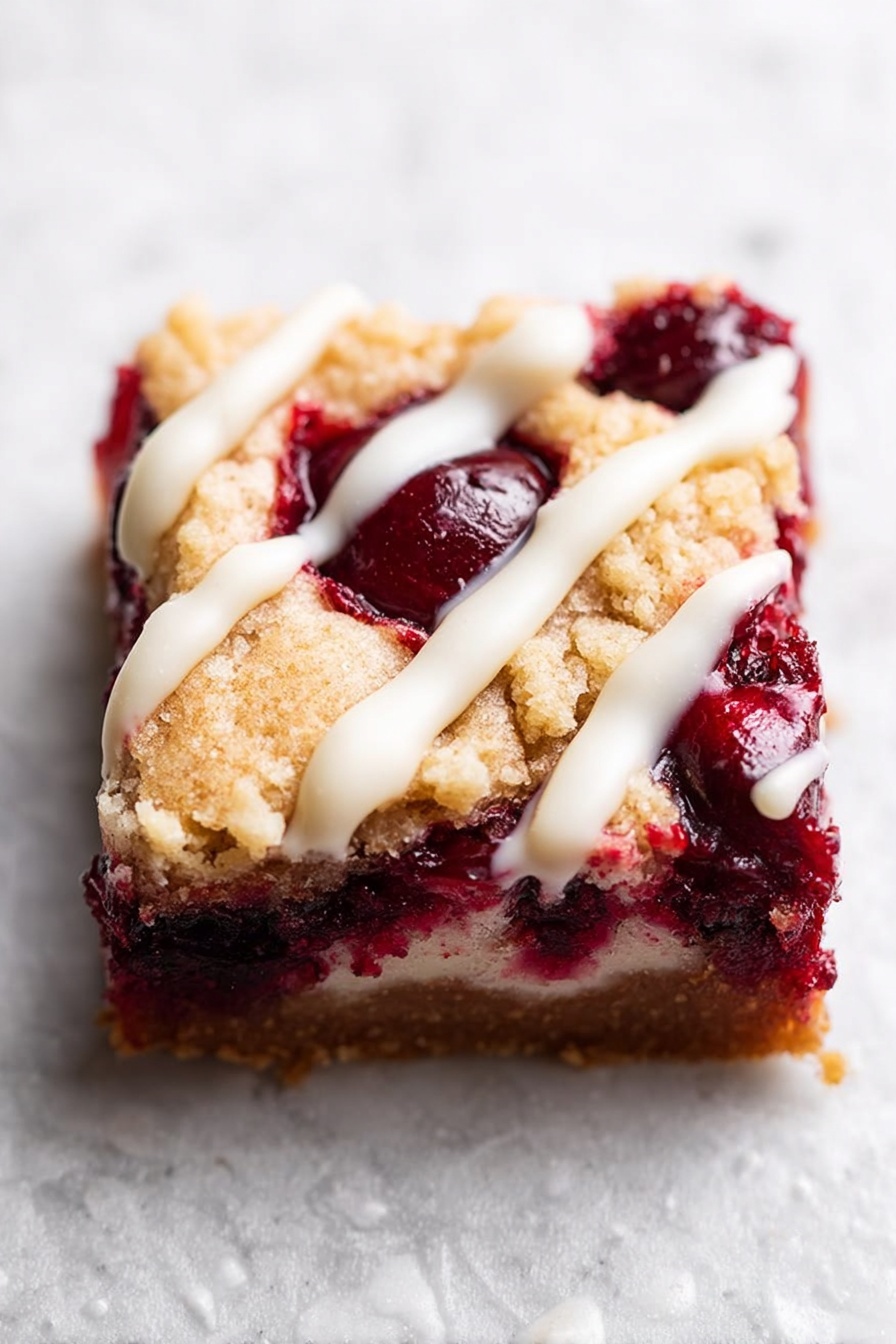 A close-up of a square slice of dessert with two visible layers: the bottom layer is thick, dark red with whole and mashed cherries visible inside, and the top layer is a golden brown, slightly crumbly crust with a rough texture. There are thin white icing drizzles on top that add a contrasting color. The dessert is sitting on a white marbled textured surface. Photo taken with an iphone --ar 2:3 --v 7