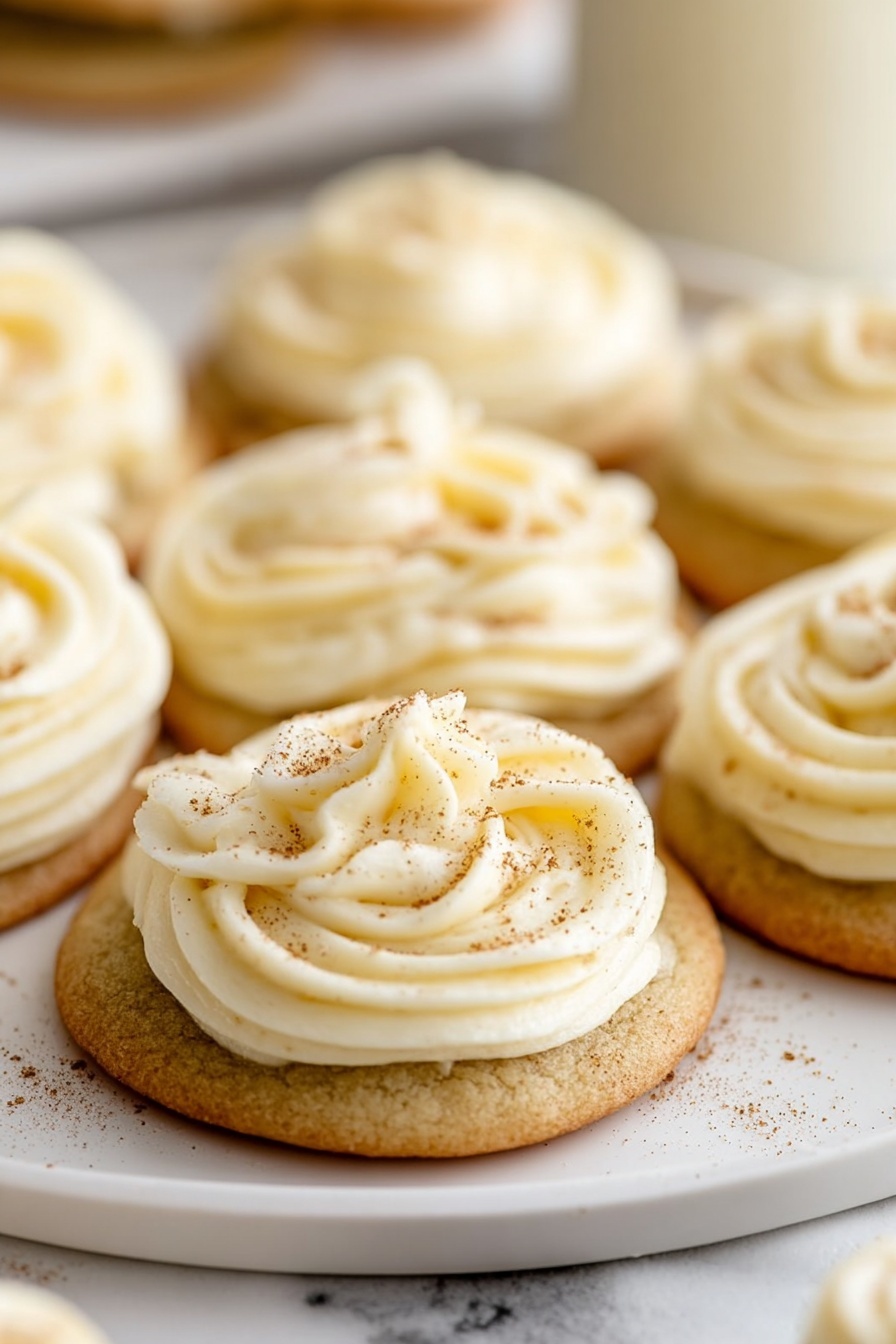 The image shows a close-up of several cookies arranged on a white plate with a white marbled surface underneath. Each cookie has one layer of light brown, round, soft-looking base with a smooth texture. On top of this base, there is a thick layer of creamy white frosting, piped in a swirling rose pattern with smooth, velvety texture and slight peaks. The frosting is sprinkled with a light dusting of brown spice powder, adding a touch of contrast. The background is softly blurred, keeping the focus on the cookies in the foreground. Photo taken with an iphone --ar 2:3 --v 7