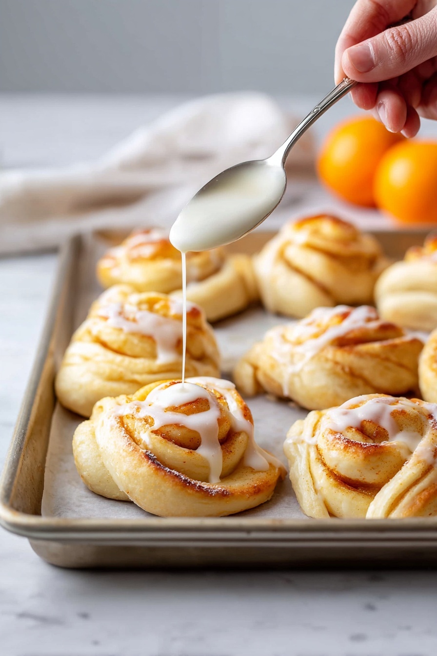 A tray of eight golden-brown twisted buns sits on a silver baking sheet with a white marbled surface below, each bun showing layers of soft dough spiraled together with a slightly browned outer crust. A woman's hand holds a spoon above the closest bun, pouring a smooth, white glaze that drizzles slowly onto the top swirl, creating a glossy drop that contrasts with the textured baked dough. In the background, two bright orange fruits add a pop of vibrant color against the light, soft-focus backdrop. photo taken with an iphone --ar 2:3 --v 7
