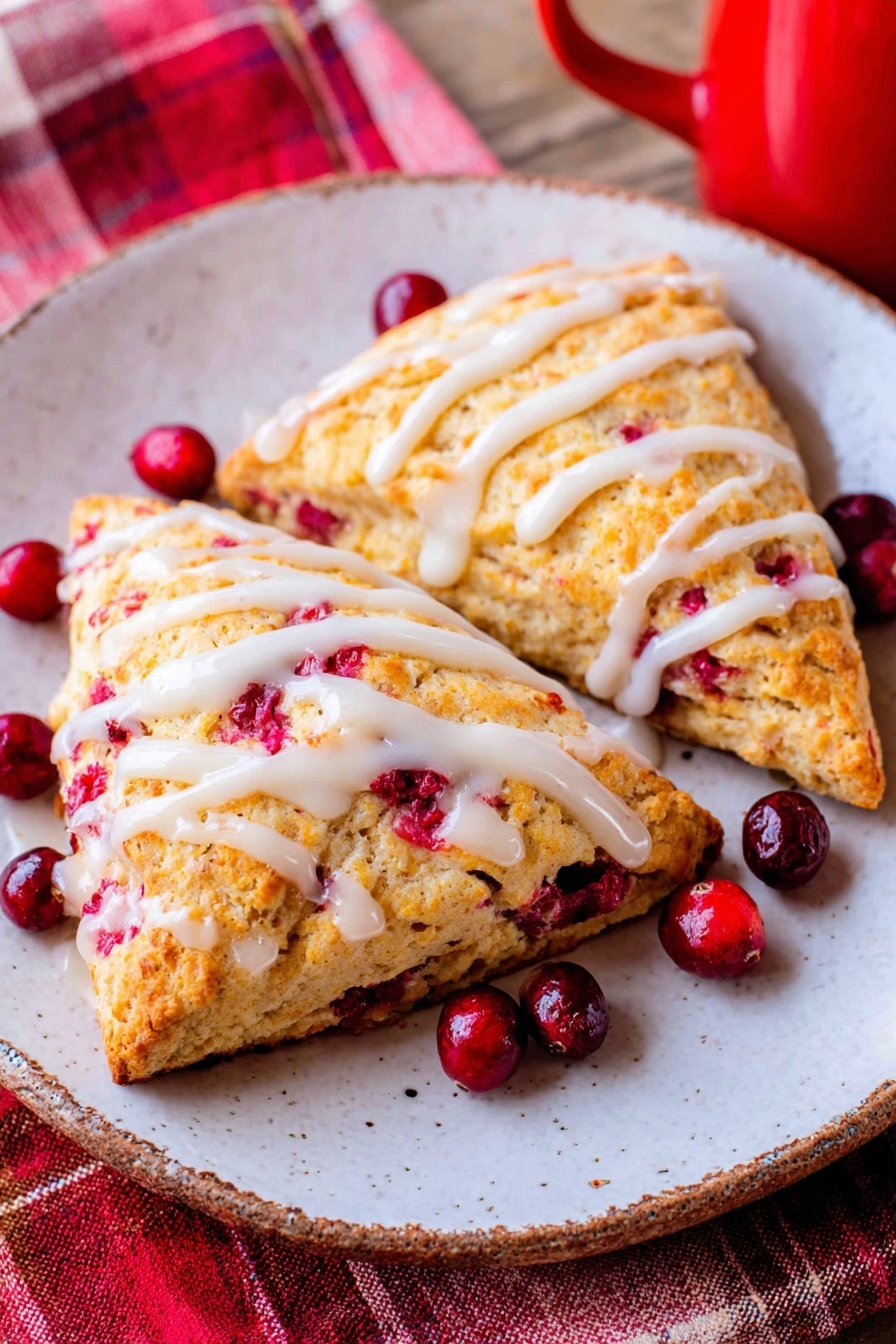 Two golden brown triangular scones sit side by side on a white plate with a white marbled texture underneath. The scones have a rough, crumbly texture with bright red berry pieces visible inside and on top. Thin white icing is drizzled in uneven lines across the top of each scone. Around the scones are a few whole fresh red berries. A red and white checkered cloth is partly visible near the bottom of the image. photo taken with an iphone --ar 2:3 --v 7