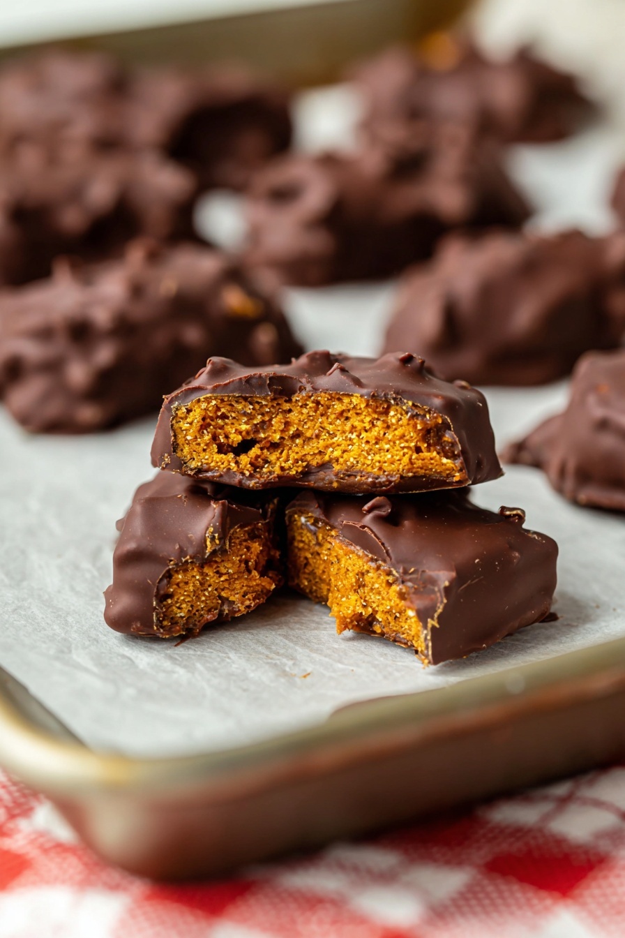 The image shows chocolate-covered treats on a baking sheet with parchment paper. The main focus is on one piece broken in half, revealing three layers: a rough orange-colored inside, a thick dark brown layer of chocolate coating, and a thin crunchy outer shell around the orange part. The broken treat pieces have a slightly bumpy texture and the chocolate looks smooth and shiny. Around the main piece, there are more chocolate-covered treats, some blurry in the background. The surface under the sheet has a white and red checkered cloth, on a white marbled texture. photo taken with an iphone --ar 2:3 --v 7