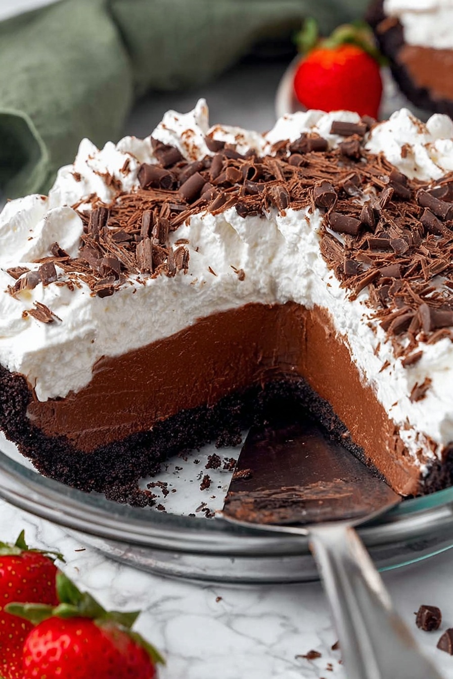 A chocolate pie with three visible layers in a round, clear glass dish on a white marbled surface. The bottom layer is a dark, moist chocolate crust that holds the pie together. Above that is a thick, rich chocolate filling with a smooth and creamy texture. The top layer is a thick, fluffy white whipped topping that covers the whole pie. Dark chocolate shavings and small chocolate curls are scattered generously over the whipped layer. A slice of the pie is being lifted by a metal spatula, showing all three layers clearly. Nearby on the surface are halved strawberries and more chocolate shavings. Photo taken with an iphone --ar 2:3 --v 7