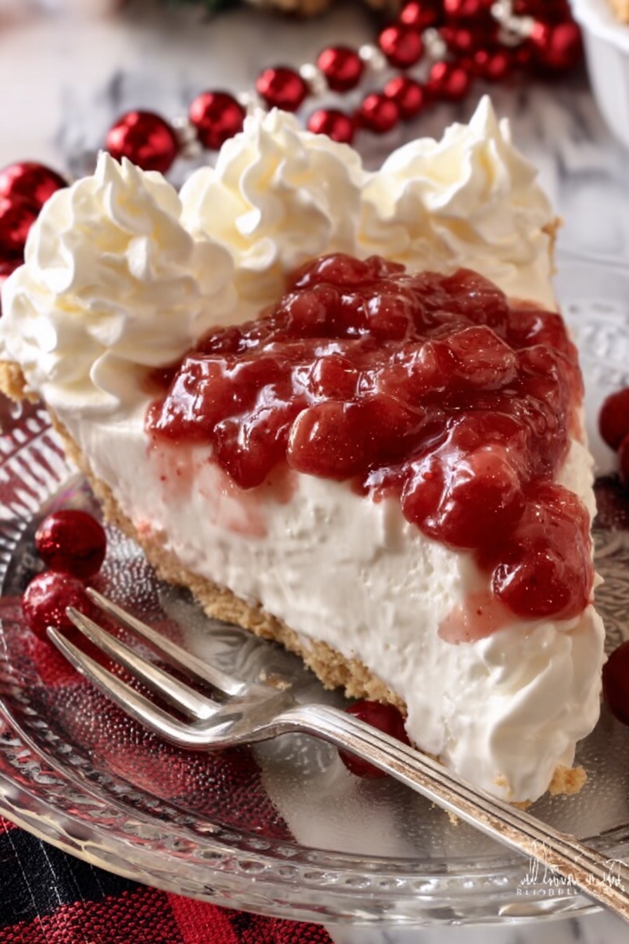 A clear white glass plate holds a slice of pie with three main layers. The bottom layer is a light brown crust that looks firm and crumbly. Above the crust is the thick white creamy middle layer, smooth and soft. On top, there is a layer of red fruit sauce with chunks, which spills slightly over the edge. Around the pie slice, dollops of whipped cream with a soft, fluffy texture create a border. The plate is placed on a white marbled surface decorated with red and black holiday-themed cloth and shiny red beads. A silver fork rests on the plate near the pie slice. Photo taken with an iphone --ar 2:3 --v 7