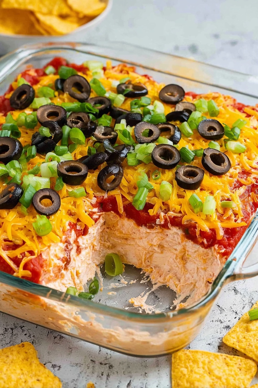 The image shows a clear square glass dish with a colorful layered dip inside. The bottom layer is creamy and light brown, topped by a thick bright red tomato or salsa layer. Above that is a large, even layer of shredded yellow cheddar cheese. On top of the cheese, there are chopped green onions and sliced black olives scattered evenly. A scoop has been taken out from the corner of the dish, showing all the layers clearly. Around the dish, there are some yellow corn tortilla chips on a white marbled surface. photo taken with an iphone --ar 2:3 --v 7