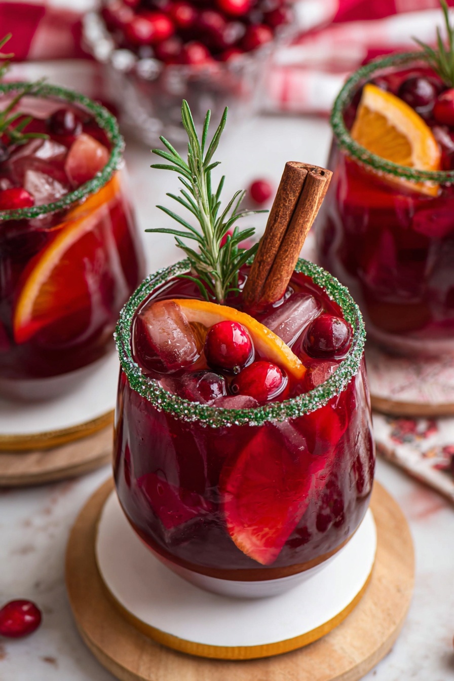 The image shows three glasses filled with a deep red drink. Each glass has a green sugar rim and is filled with ice cubes, floating cranberries, and slices of orange and red fruit. One glass, in focus, has a cinnamon stick and a rosemary sprig placed inside, standing upright. The glasses are clear and set on round white coasters with gold rims, placed on a light wooden board. The background has a white marbled texture with a blurred red and white cloth and extra cranberries scattered around. photo taken with an iphone --ar 2:3 --v 7
