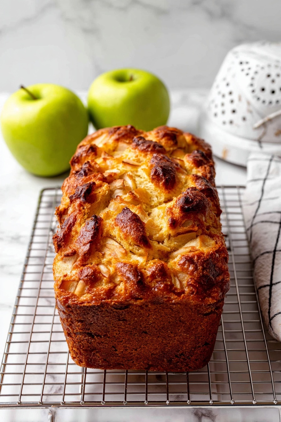 This image shows a single loaf of apple bread with a rough, uneven top layer that is golden brown with some darker brown spots, giving it a baked, crispy texture. The loaf is placed horizontally on a metal cooling rack with a shiny, grid pattern, resting on a white marbled surface. To the left and right of the bread, there are two whole green apples. At the back on the right side, there is a white colander with small round holes, and a white and black checkered cloth is partially visible in the background. The photo taken with an iphone --ar 2:3 --v 7