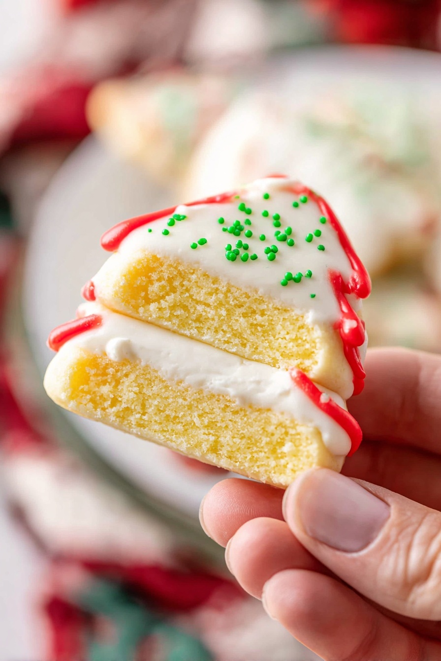 A close-up view of a small, square-shaped cake piece held by a woman's hand showing two layers of soft yellow sponge with a thick layer of white cream filling in the middle. The cake is fully covered in a smooth white icing with a thin layer of shiny red icing on the top edge. Few tiny green sprinkles are visible on the white icing around the sides. The background features a white marbled texture with blurred hints of red, white, and green colors. Photo taken with an iphone --ar 2:3 --v 7