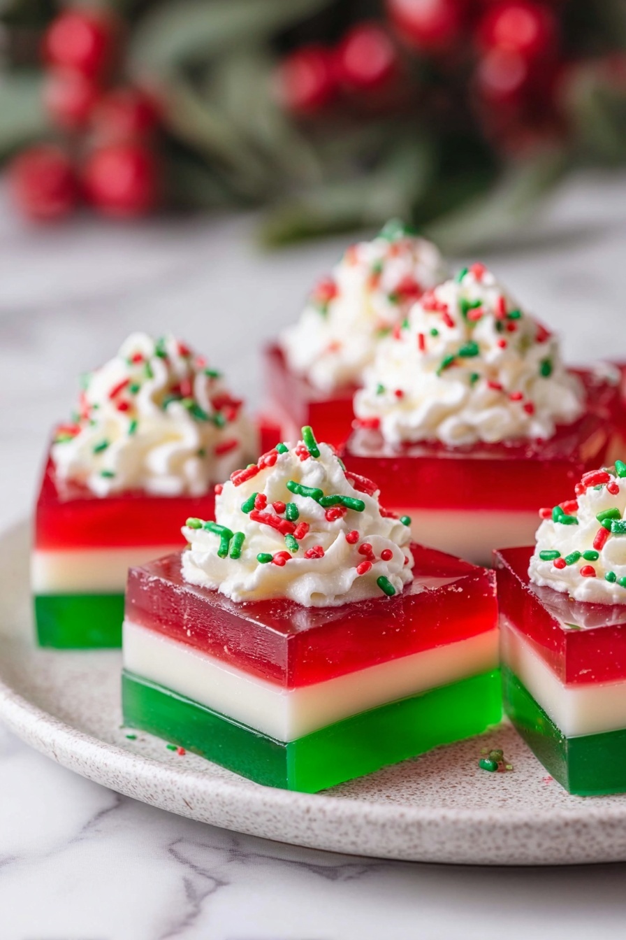 The image shows several square jello desserts arranged on a white plate with a white marbled surface underneath. Each dessert has three clear layers: a glossy green layer at the bottom, a creamy pale yellow layer in the middle, and a bright red translucent layer on top. Each piece is topped with a dollop of white whipped cream and decorated with small red and green sprinkles. In the background, there is a blurred festive basket with pinecones, green leaves, and red berries. Photo taken with an iphone --ar 2:3 --v 7