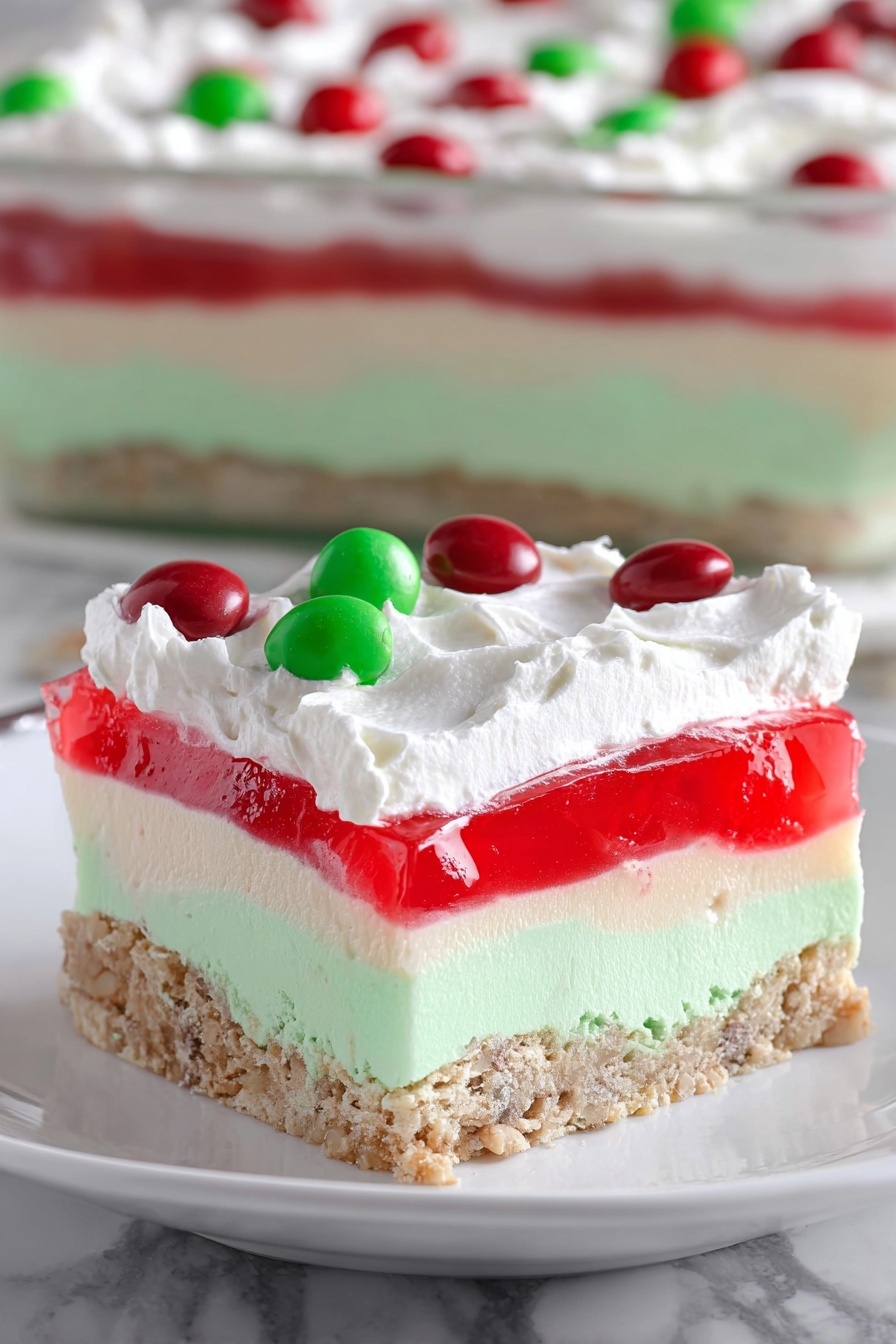 The image shows a thick square slice of layered dessert on a white plate, resting on a white marbled surface. The bottom layer is a light brown, crumbly crust with visible small nuts or grains. Above that is a thick, smooth, and light green layer with a slightly porous texture. Next is a white creamy layer that looks soft and fluffy. On top of this is a bright red, translucent gelatin layer. The dessert is finished with a thick, white whipped cream layer spread unevenly on top. Scattered on the whipped cream are shiny, round candies in red and green colors. In the background, a larger, rectangular version of the dessert is visible in a clear dish. photo taken with an iphone --ar 2:3 --v 7