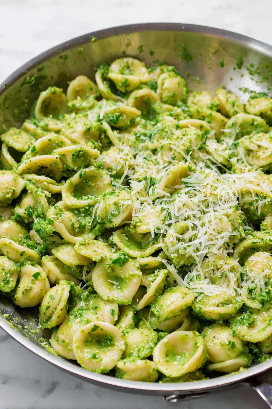 A close-up view of a large silver pan filled with orecchiette pasta coated evenly in a bright green pesto sauce. The small, round ear-shaped pasta pieces give a textured look, each piece covered in tiny bits of green herbs and oil from the pesto. On top, a light sprinkle of grated pale yellow cheese adds a soft contrast against the green pasta. The pan sits on a white marbled surface, and the photo captures the fresh and creamy texture of the sauce along with the hearty pasta pieces. photo taken with an iphone --ar 2:3 --v 7