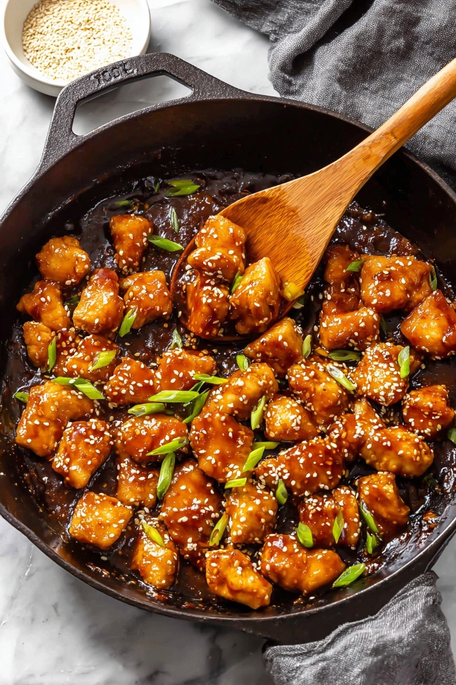 A black cast iron skillet filled with small, golden-brown chicken pieces cooked in a shiny, dark brown sauce with a slightly sticky texture, topped with white sesame seeds and bright green chopped scallions scattered evenly on top and between the chicken pieces; a wooden spoon resting inside the skillet holds some chicken, showing the sauce’s glossy surface underneath. The skillet is placed on a white marbled surface with a gray cloth slightly visible in the background, and a small white bowl with sesame seeds sits nearby. photo taken with an iphone --ar 2:3 --v 7