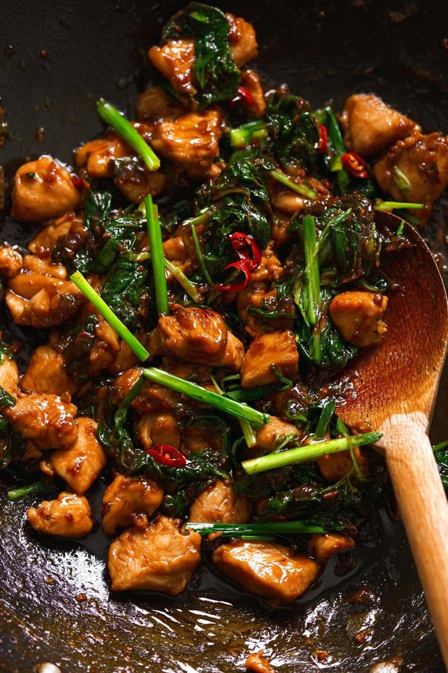 The image shows a close-up of a dark wok filled with small, glossy pieces of brown cooked chicken mixed with dark green leafy vegetables and thin slices of bright green scallions. The chicken pieces have a smooth, shiny texture, coated in a rich sauce with small red chili flakes scattered throughout. A wooden spoon is partially visible stirring the contents on the right side, with the handle extending out of the frame. The wok surface shows some oil reflections and seasoning spots. Photo taken with an iphone --ar 2:3 --v 7
