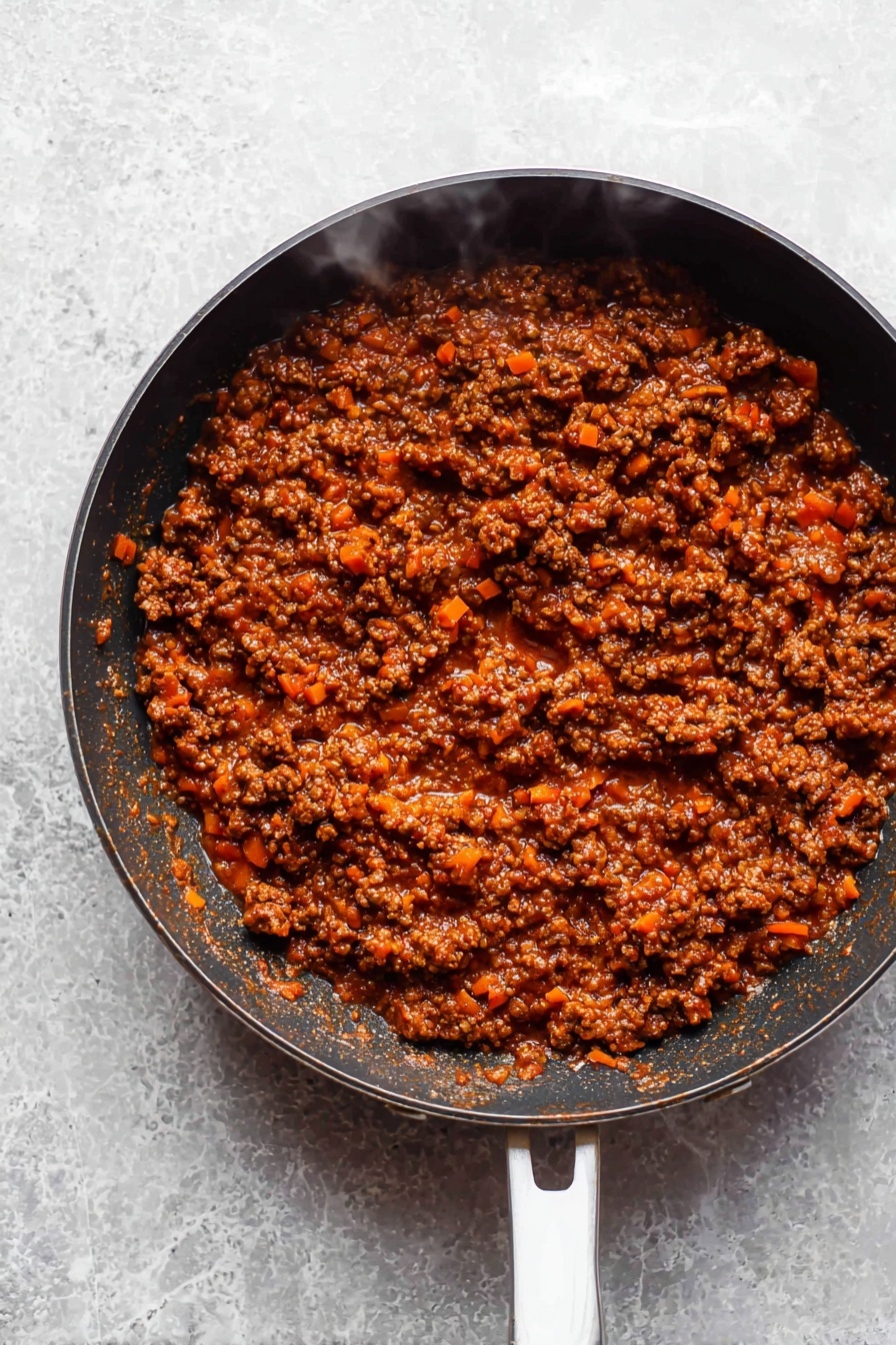 The image shows a black frying pan filled with a thick, rich meat sauce. The sauce has small pieces of browned ground meat mixed with finely chopped orange carrots, all coated in a dark reddish-brown tomato sauce that looks chunky and hearty. The frying pan's handle is white with a slight gray mark, and the pan sits on a white marbled surface. Steam rises gently from the sauce, adding a sense of warmth and freshness to the dish. photo taken with an iphone --ar 2:3 --v 7