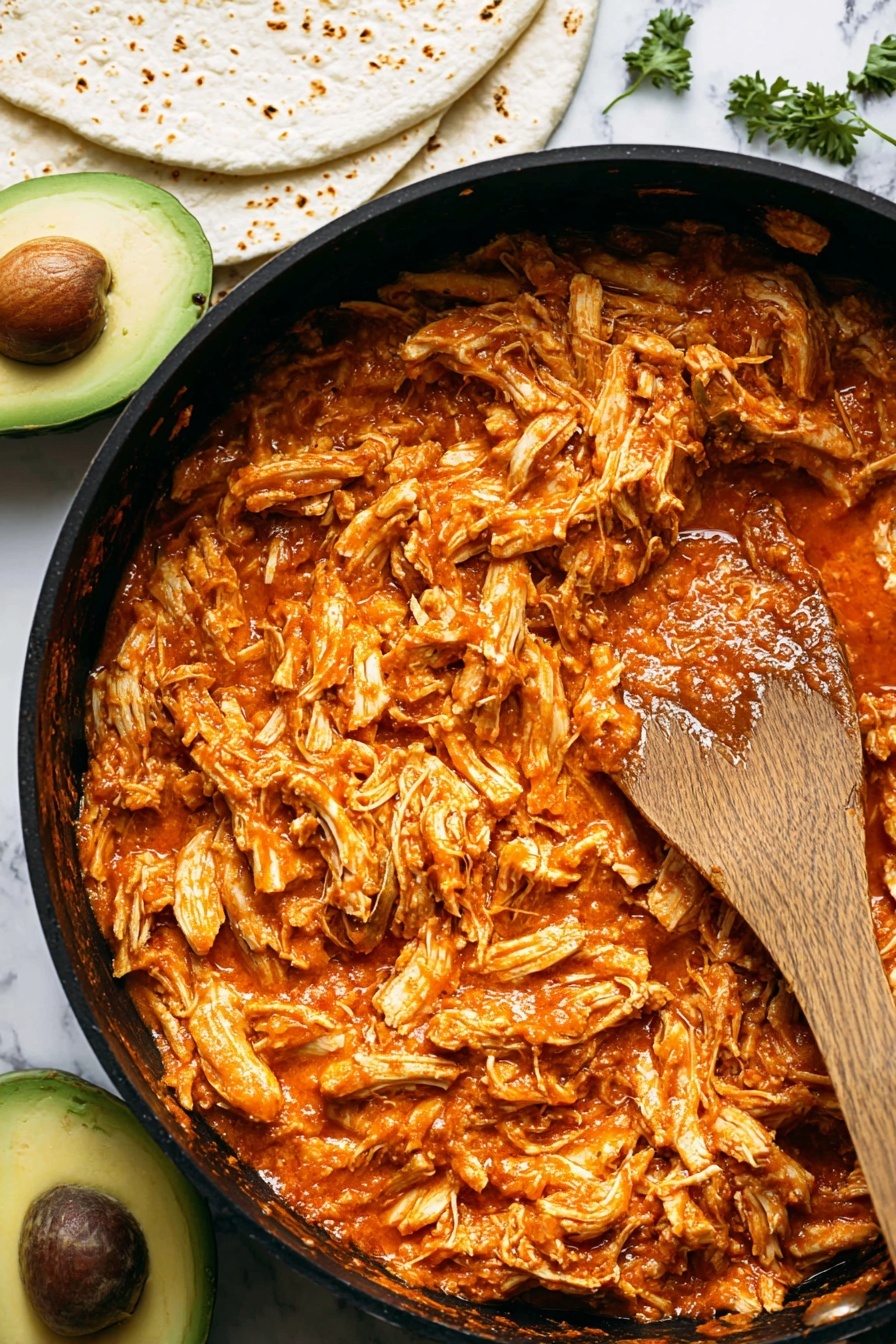 A close-up of a large black pan filled with shredded chicken mixed in a thick, orange-red sauce covering the entire pan surface, with visible juicy texture and slightly glossy finish. A wooden spoon rests on the right side of the pan, coated with some of the sauce. Below the pan, part of a sliced avocado with a dark seed and green flesh is visible, next to some white tortillas with light brown char marks. The background has a white marbled texture, and there are small green herb pieces scattered around. Photo taken with an iphone --ar 2:3 --v 7